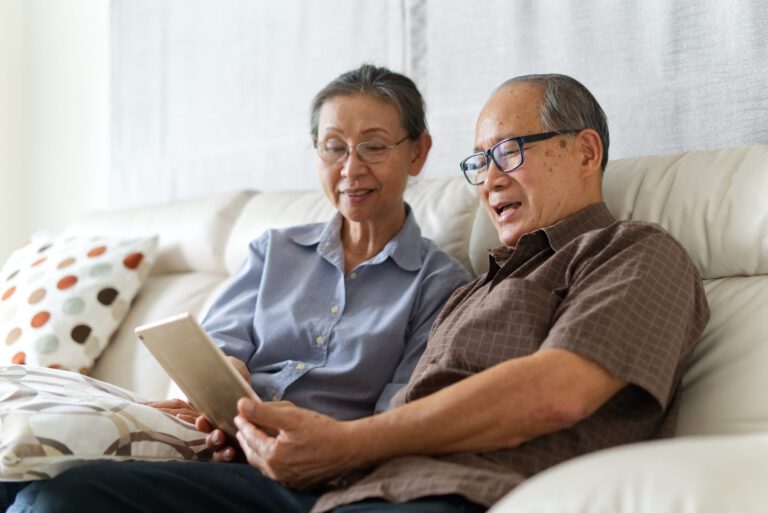 Couple senior assis sur un canapé à la maison, jouant sur une tablette et se relaxant ensemble. Ils sont souriants et aiment passer du temps ensemble avec bonheur. Concept de vie de retraite heureuse.